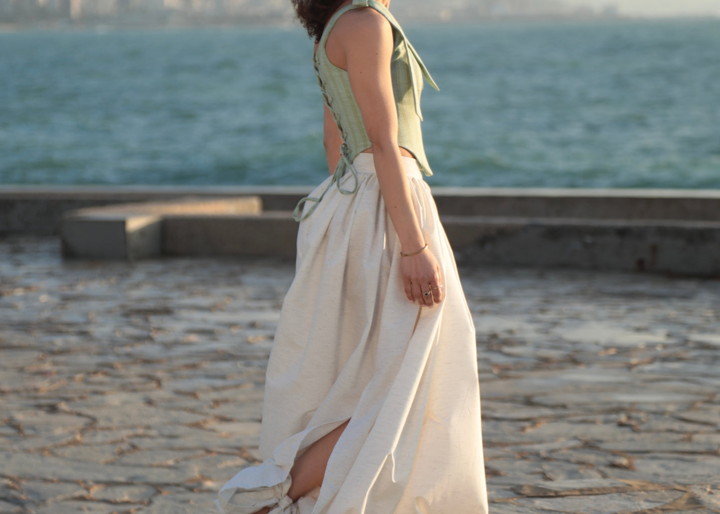 Woman in a corset and balloon linen bow trousers standing on a stone pathway by the Mediterranean.